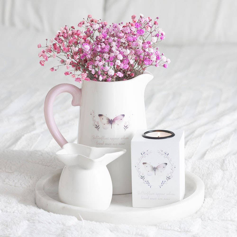 White pitcher with pink flowers and a white candle with butterfly design on a white surface