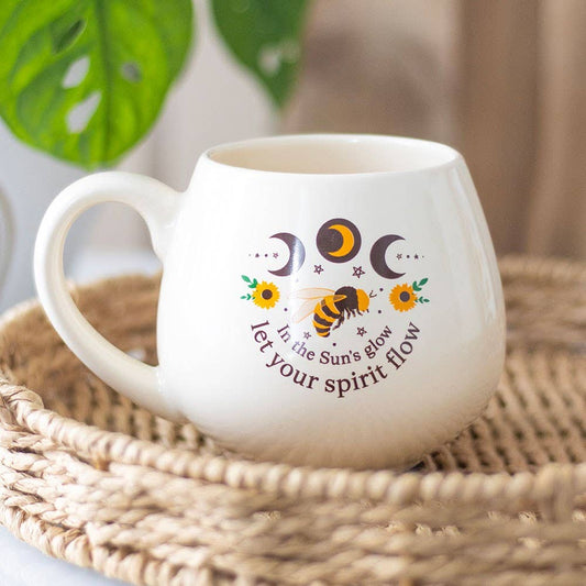 White mug with sunflower, bee, and moon phases design on a woven mat.