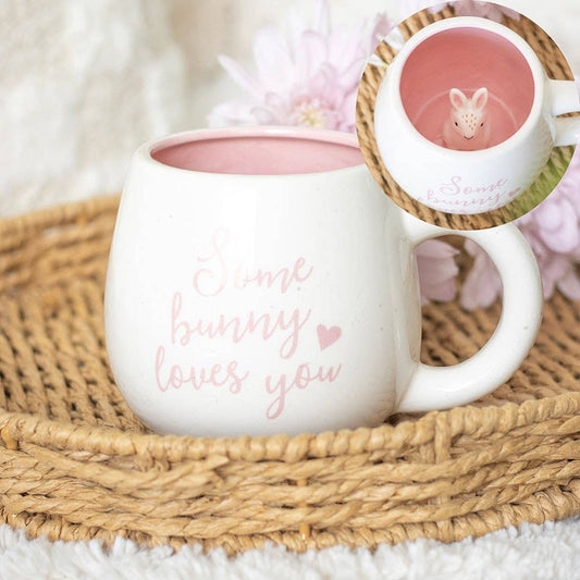 White mug with 'Some bunny loves you' text, pink interior, and small rabbit figure on a woven mat.