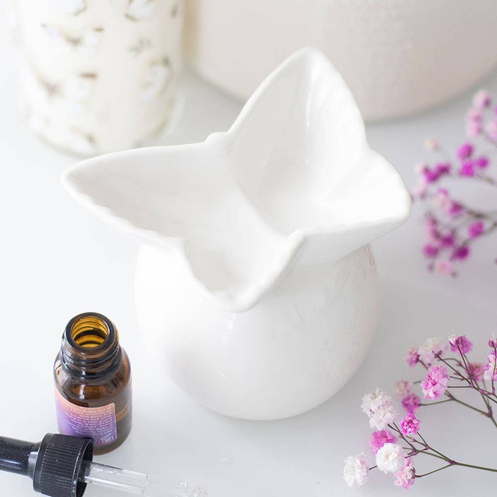 White ceramic cat-shaped diffuser with a bottle of essential oil and pink flowers on a light background