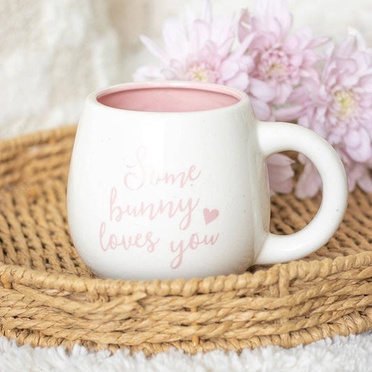 White mug with 'Some bunny loves you' text on a woven mat with pink flowers.
