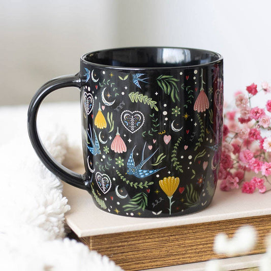 Black mug with colorful floral and heart designs on a white surface with pink flowers in the background