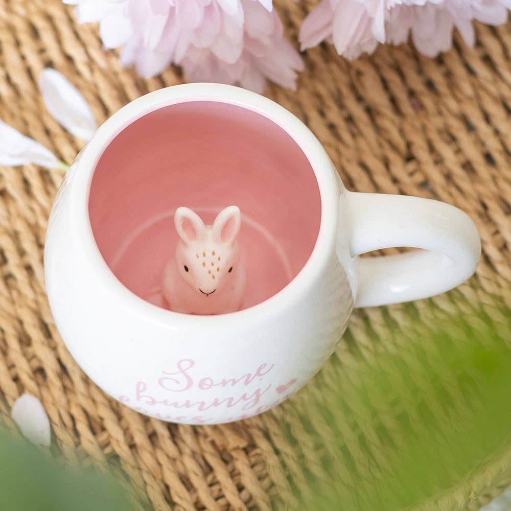 White mug with a pink interior containing a small rabbit figurine, placed on a woven mat with flowers.