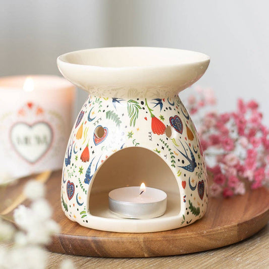 Decorative ceramic oil burner with a lit candle on a wooden tray, surrounded by flowers and a mug.